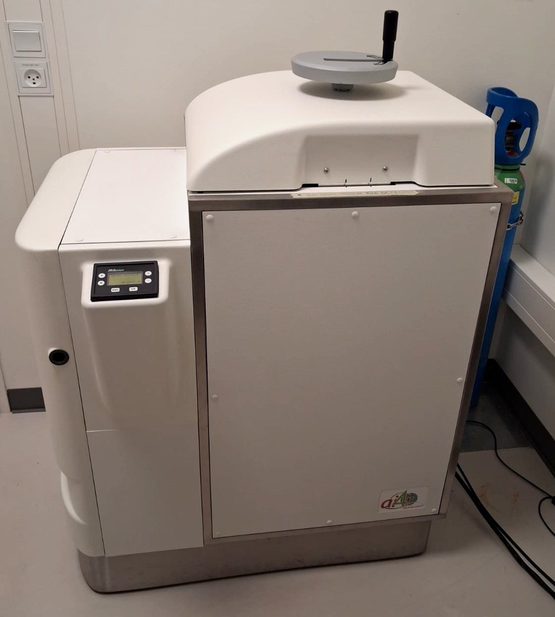 The BeaQuant seen from the front: a white cabinet with a domed top cover carrying a grey hand-wheel, a left-hand module with a small control panel and green display, and a steel-framed front panel with the ai4R BeaQuant badge at lower right. A blue gas cylinder stands behind it on the right.