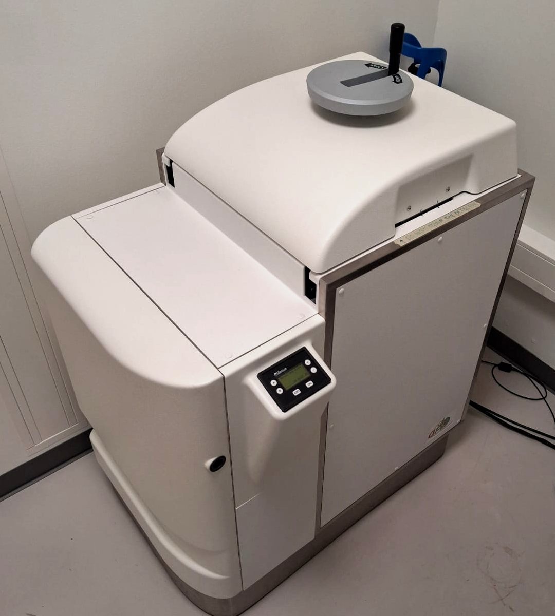 The same instrument from the side and slightly above, standing in the corner of a small white room: the rounded left-hand module with its control panel, the domed top cover with the hand-wheel, and the steel-framed front panel on a stainless-steel base.